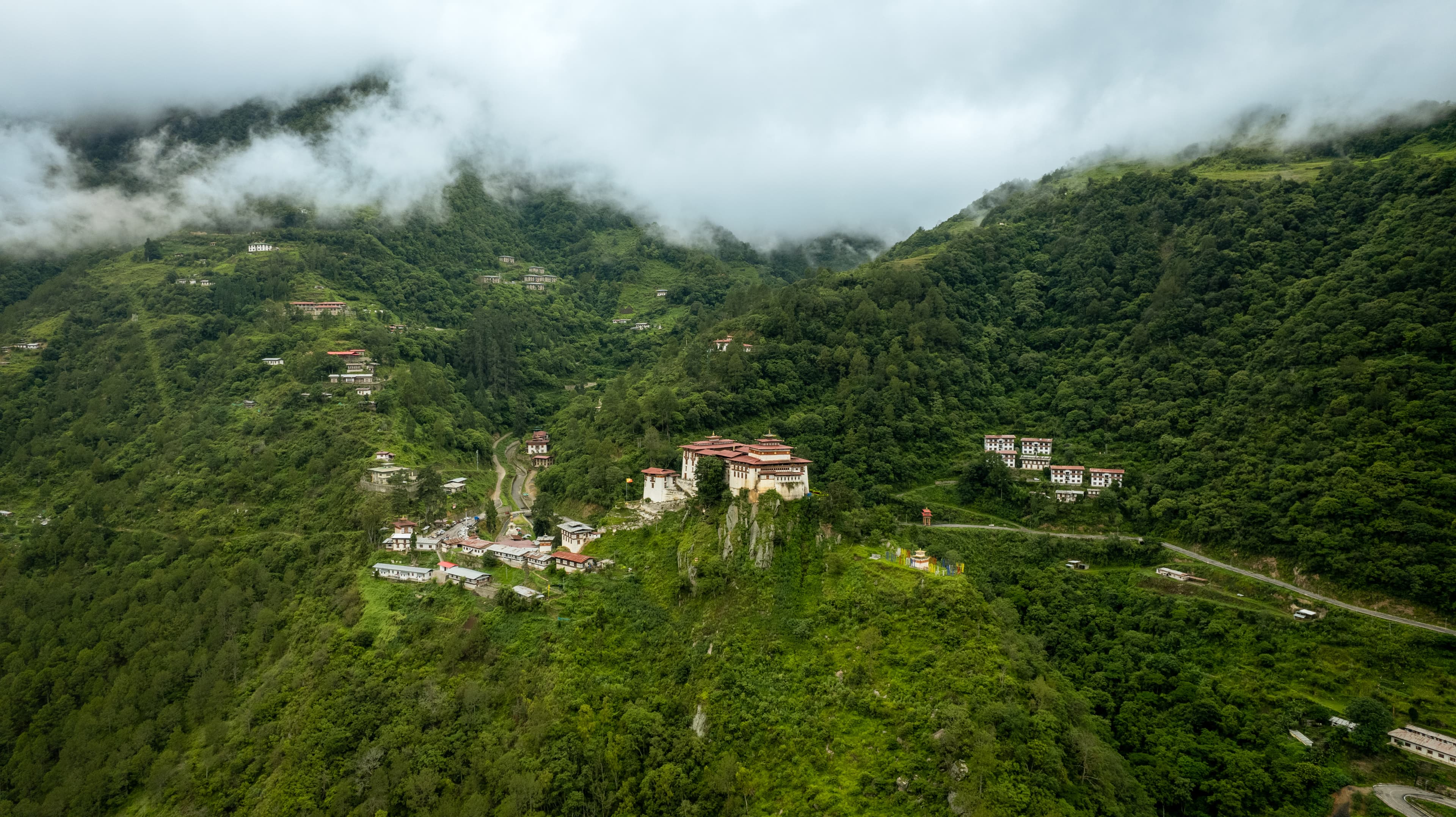 Aerial view of a Bhutanese dzong perched on a forested cliff, wreathed in cloud